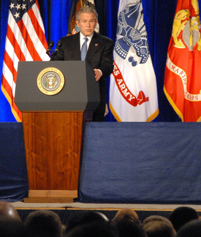 A man in a suit stands behind a lectern.  Behind him are several flags, including the U.S. flag, the Army flag, and the Marine Corps flag.