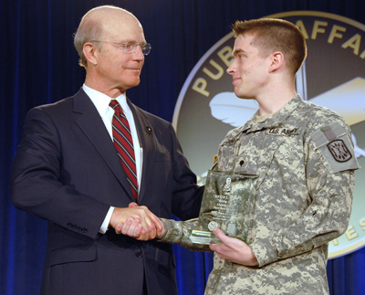 A man in a suit shakes hands with a Soldier.  The soldier holds a glass award.  Behind them is a sign that says "Public Affairs."