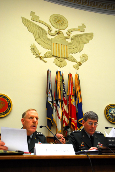 Two men in military uniforms sit at a table. One speaks into a microphone.  Behind them on the wall is a large eagle emblem.  Below that, against the wall are five flags.