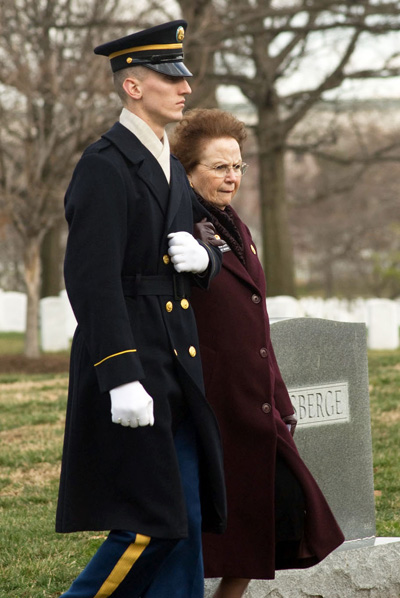 A man in a military uniform and a woman in a coat walk together through a cemetery, with their arms linked.
