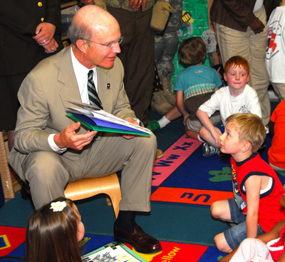 A man in civilian clothes is seated and holding several pieces of paper with drawings made by children.  A small boy seated on the floor looks up at him. Other children sit nearby.
