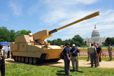 A tank-like combat vehicle sits on a dirt path near a patch of grass.  In the background is the U.S. Capitol Building.  People mill around, talking in small groups.