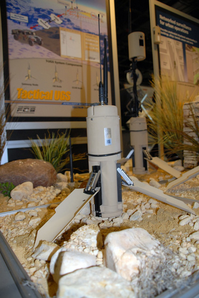 Two electronic devices that look like cylinders on tripods are situated in a box filled with soil and rocks.  Behind them on the wall is a sign that says "Tactical UGS." 