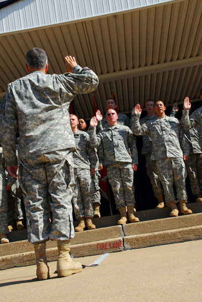 Dozens of military personnel stand under an overhang.  They have their right hands raised. In the front, a man in a military uniform faces them and also has his right hand raised.