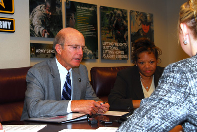 A man and a woman sit near each other at a table.  Another woman sits across from the man.  On the wall behind the man are signs illustrating soldering, and a U.S. Army logo.