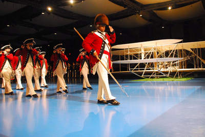 About ten individuals in Revolutionary War-era military uniforms march in formation.  Behind them is a replica of an early aircraft.