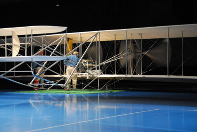 In a large room and on a shiny blue floor is a replica of an early aircraft.  A man stands at the center of the aircraft.
