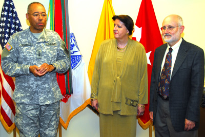 A man in a military uniform stands next to a woman and a man in civilian clothes.  Behind them are an array of flags, including the U.S. flag and the U.S. Army flag.