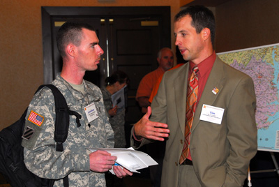 A man in a military uniform talks to a man in a suit.  Behind them is a map.