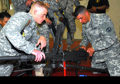 Two men in military uniforms kneel on the ground and perform maintenance operations on a machine gun.