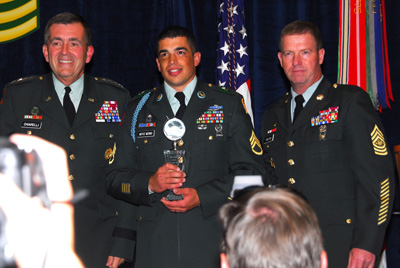 Three men in military uniforms stand near each other.  One holds a trophy.  Behind them is an American flag.