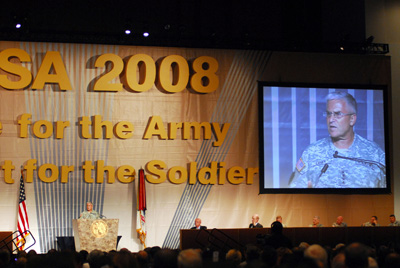 At the far front of a cavernous convention center room, a man in a military uniform stands behind a lectern.  He also appears in close-up on a projection screen at the right side of the room. Also on the stage, about seven people sit at a long table.  A crowd faces the stage.