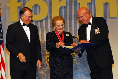 A woman is flanked by two men in suits.  She has a ribbon around her neck.  She and one of the men are looking at a document in a folder.