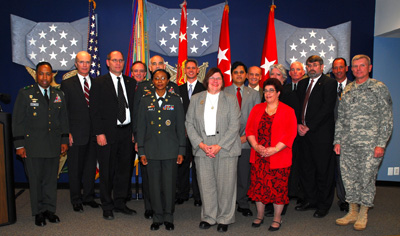 More than a dozen people, in both civilian clothing and military uniforms stand together in front of flags and wall hangings that represent various versions of the Medal of Honor. 