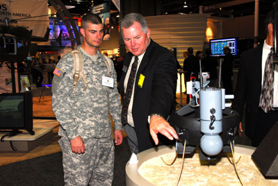 A man in a military uniform stands next to a man in a suit.  They look at an aerial drone on a table. n the background are a variety of other displays and signage.