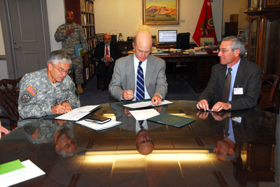 A man in a military uniform and two civilian men sit at a table.  Two of the men sign documents.
