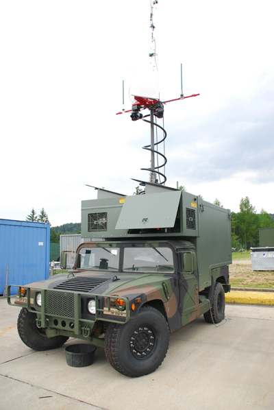 A green, boxy military vehicle has a large antenna on top.