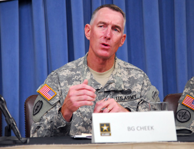 A man in a military uniform sits at a table. In front of hims is a sign that says "BG Cheek." behind him are blue drapes.