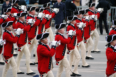 More than a dozen military musicians in red, Revolutionary War-era uniforms, march in a parade.