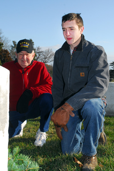 A young man is on one knee in front of a white tombstone.  Near him is an older man, also on one knee, wearing a ball cap that says "Korea Veteran." They have placed a wreath at the tombstone.