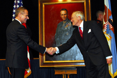 Two men in suits shake hands.  Behind them is a painting that features one of the two men, but in a military uniform.  There are flags on either side of the painting.