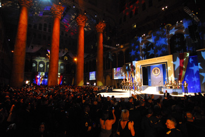 Pillars lit up with red lights are on the left side of a huge, darkened room. At the front of the room is a stage, lit up with spotlights.  A crowd is gathered in front of the stage. Words above the stage say "Renewing America's Promise."  Soldiers stand on the stage in formation.