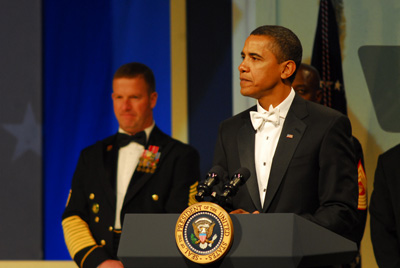 A man in a tuxedo stands behind a lectern. A symbol on the lectern says "Seal of the President of the United States."  Behind him are several men in military uniforms.