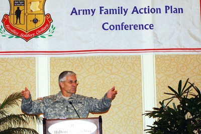 A man in a military uniform stands behind a lectern.  His arms are outstretched.  Above him a banner near the ceiling says "Army Family Action Plan Conference."