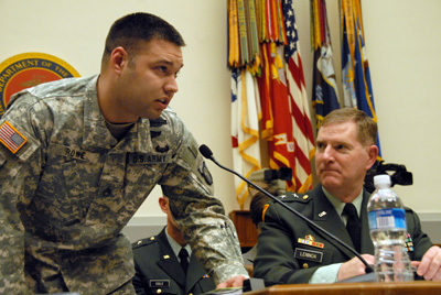 A man in a military uniform stands and leans into a microphone.  Nearby, another man in a military uniform is seated and watches him.  Multiple flags representing the U.S. military services are at the back of the room.