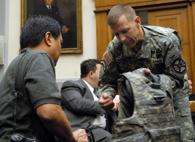In a large room, a man in a military uniform holds a body armor vest.  A civilian man observes. There is a painting on the wall behind them.