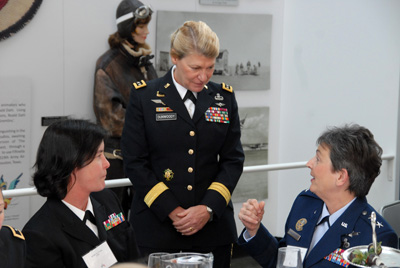 A woman in a military uniform stands and speaks to other women in military uniforms who are seated at a table. Behind them is what appears to be a display that features photos and words on the wall, and a female mannequin in a military uniform.