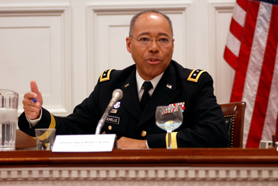 A man in a military uniform sits at a table and speaks into a microphone.  Behind him is an American flag and wainscoting.  