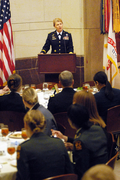 A woman in a military uniform stands behind a lectern at the front of a room where people are seated at round tables and eating.  To her right is an American flag, and to her left is the U.S. Army flag.