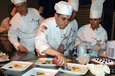 Three individuals in chef's outfits watch a fourth use a squeeze bottle to apply fruit coulis to a plate, which already contains a slice of pie. Other plates and deserts are nearby.