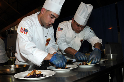 Two men in chefs outfits place food on white plates.  They wear blue rubber gloves.  