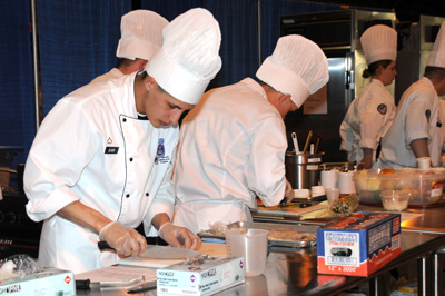 A man in a chef outfit uses a knife to cut fish.  Others in chefs outfits are near him doing similar tasks.
