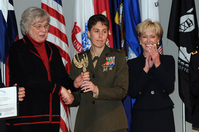 A woman in a military uniform holds a trophy.  She is flanked by two women in civilian clothes.  Behind them is an array of flags, including the U.S. flag, the Army flag, the Air Force flag, and the POW/MIA flag.