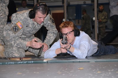 A civilian man lies on the ground and points a rifle downrange.  Next to him, seated on the floor, is a soldier.  