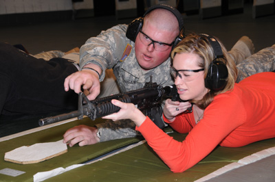 A woman in a red shit lays on the ground and aims a rifle.  A man in a military uniform lays next to her and points at the sights on the weapon. Both wear ear and eye protection.