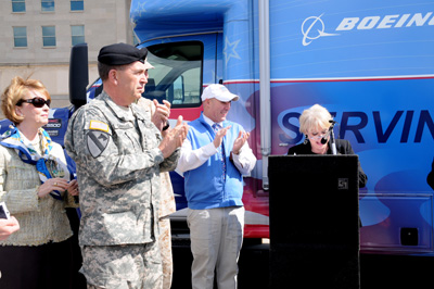 Outdoors, a civilian woman stands behind a lectern. Nearby are other civilians and a man in a military uniform, who wears a black beret.  Behind them is a large vehicle with the word "Boeing" printed on the side.  In the far background in the Pentagon.