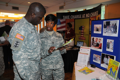 A man and a woman in military uniforms stand in front of a table with paperwork and cardboard displays.  In the background is another display that says "Take Charge at No Charge" and also "Military One Source."