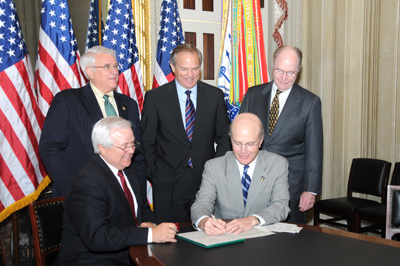 Two men in suits are seated at a table and appear to be involved in signing something together.  Three men stand behind them.  An array of flags are behind them, including at least four American flags and one U.S. Army flag.