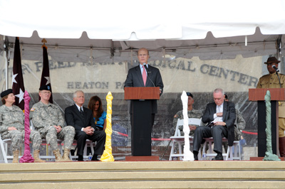 A man in a suit stands behind a lectern on a stage.  Behind him, words on the side of a building say "Walter Reed Army Medical Center - Heaton Pavilion."  Soldiers and other civilians are seated on the stage.