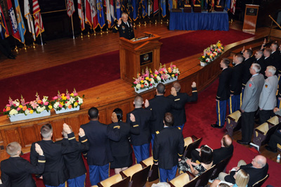 In a large auditorium with a wooden stage, dozens of uniformed soldiers stand in front of wooden chairs and raise their right hand.  On the stage a man in a military uniform stands behind a lectern. his right hand is raised.  Behind him on the stage are more than a dozen flags. In front of him are flowers in boxes.