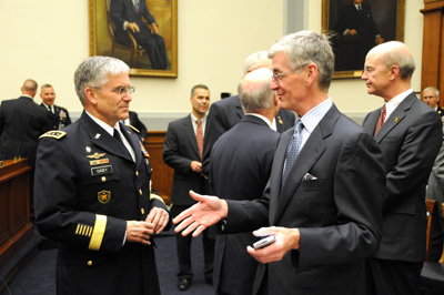 In a large room, a man in a military uniform talks with a man in a suit.  Nearby, other men in suits talk in a small group.  Paintings hang on the wall.