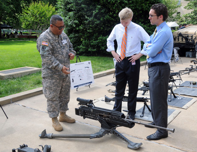 In an outdoor area, multiple guns sit on a concrete patio.  A soldier and two civilian men stand around a machine gun which is mounted on a tripod.  A sign in the background provides details on the weapons.