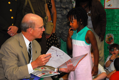 A man in civilian clothes is seated and holding several pieces of paper with drawings made by children.  A small girls talks with him.  She has her finger pressed against her teeth. Other children sit on the floor near the man.  Adults stand in the background.      
