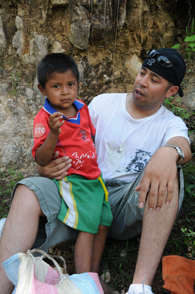 A man sits and has his arm around a young boy in a red shirt.  Cloth bags are at their feet.