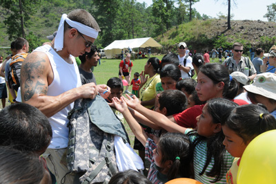 A young man wearing a head band stands amidst a crowd of people, including children, and appears to be handing something out to them.  They reach out to him with their hands outstretched. 