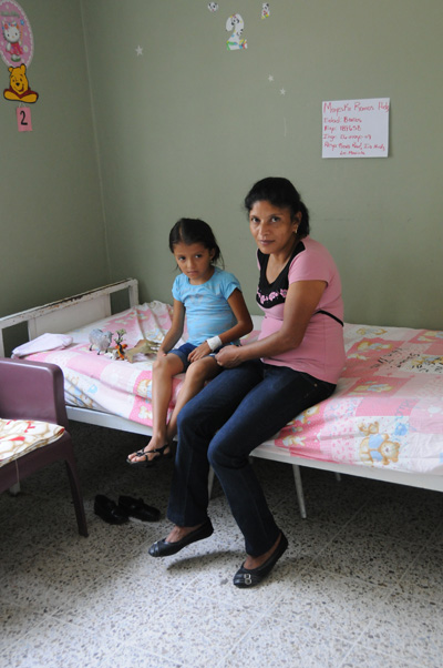 A young girl and a woman sit on a bed, adorned with pink bed sheets.  A Winnie the Pooh sticker is on the wall above the head of the bead.  On the adjoining wall is a handwritten sign with information written in Spanish. Some small toys are also on the bed.  